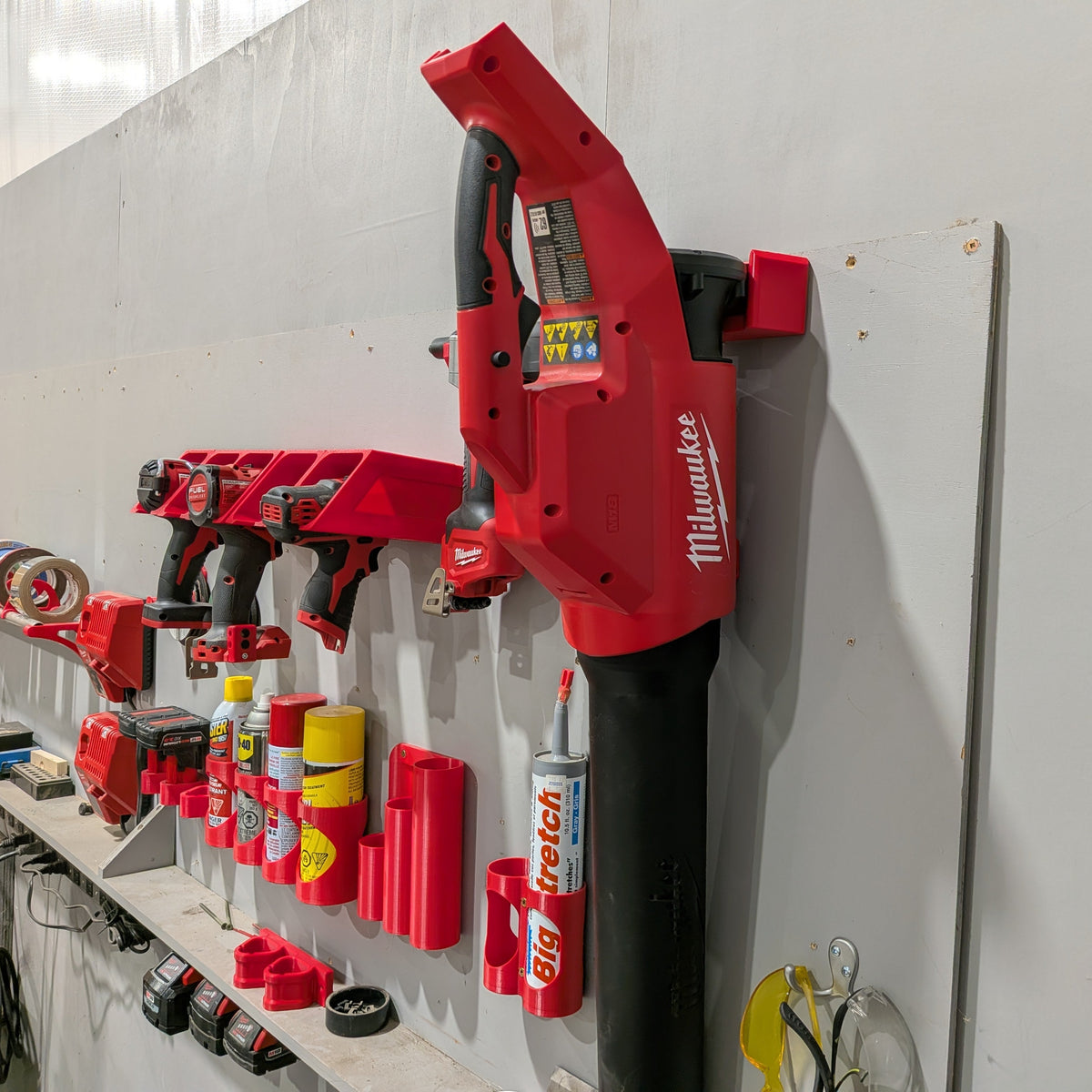 A red Milwaukee cordless leaf blower mounted on a white wall next to a workbench holding various power tools and accessories.