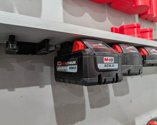 Three black and red M18 cordless tool batteries mounted on a white wall shelf, with red storage bins visible in the background.