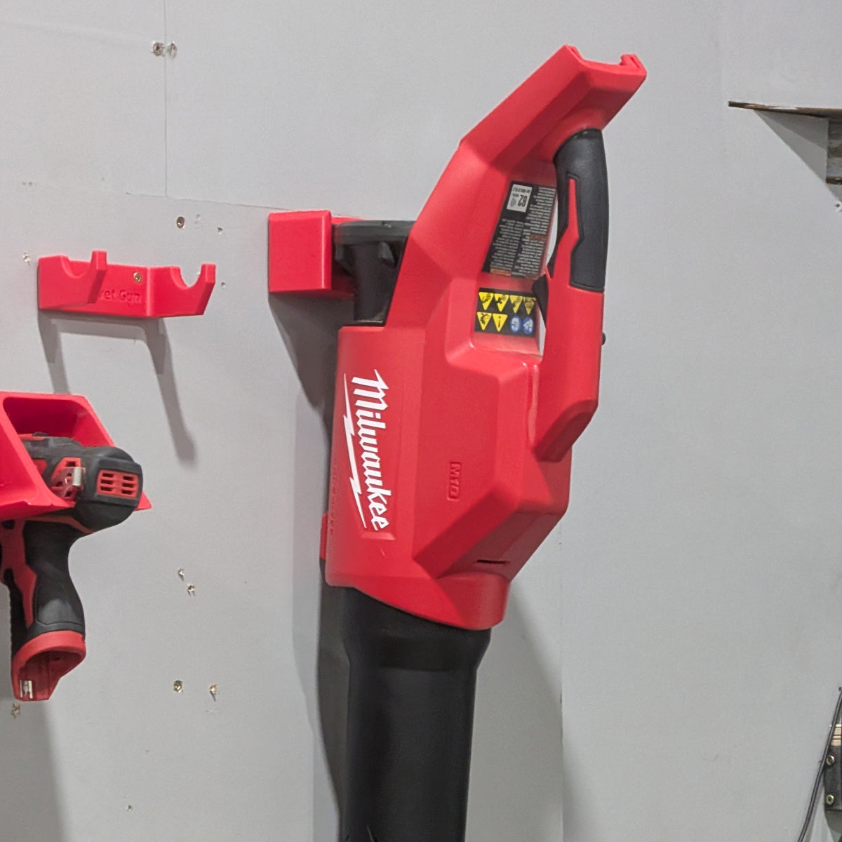 Milwaukee cordless leaf blower in red and black hanging on a gray workshop wall next to other Milwaukee tools.