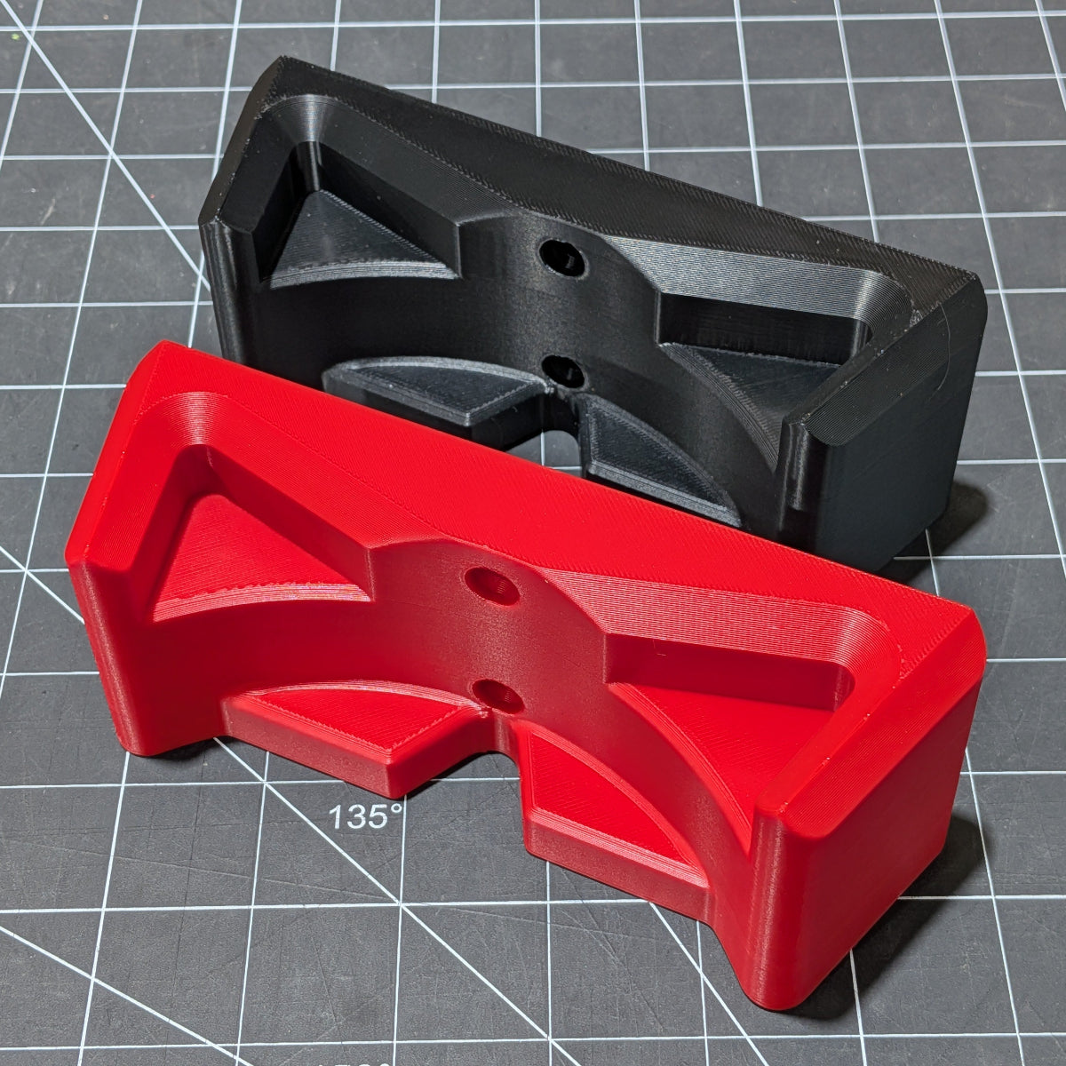Two 3D-printed plastic components, one in red and one in black, displayed on a gray cutting mat with grid lines. The red piece is in the foreground, and the black piece is positioned behind it at an angle.