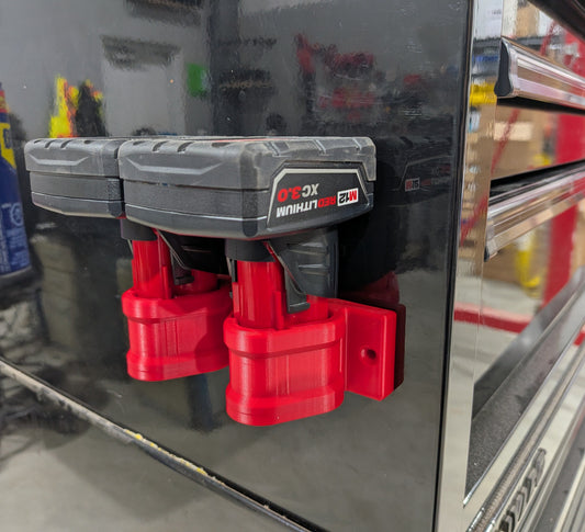 Two Milwaukee M12 batteries with black bodies and bright red mounting brackets attached magnetically to a glossy black tool chest. 