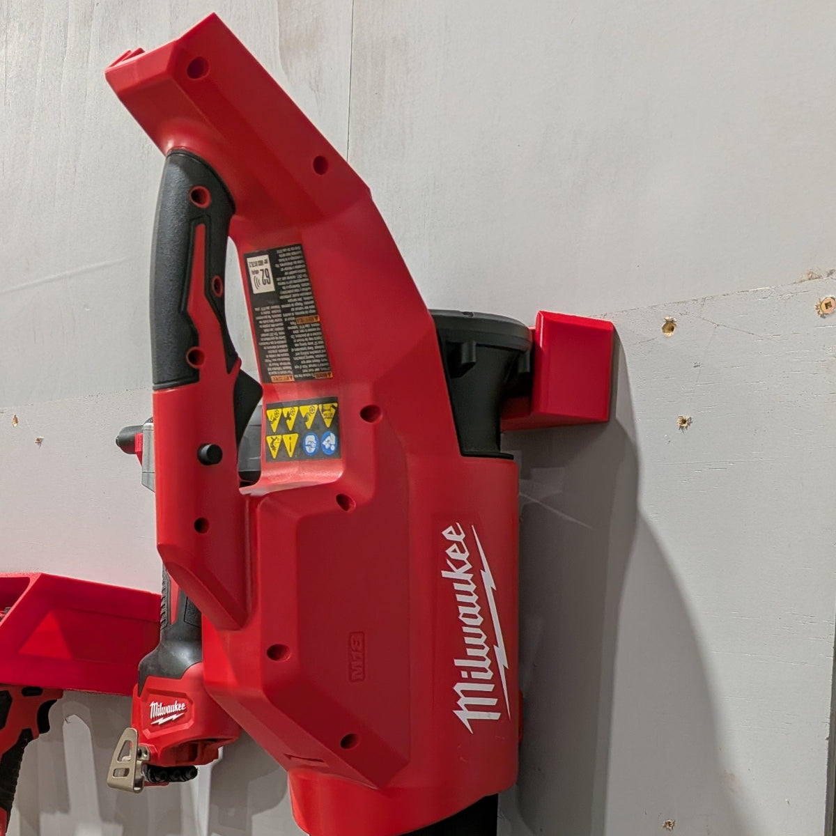 A red Milwaukee cordless leaf blower mounted on a white wall next to a workbench holding various power tools and accessories.