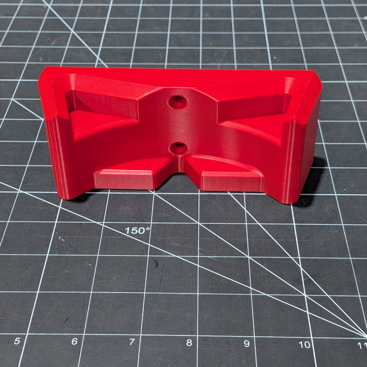 A bright red 3D-printed plastic M18 Blower Hanger with a symmetrical design and two circular holes, placed on a black cutting mat with grid lines.