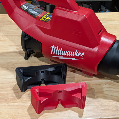 Milwaukee red and black cordless leaf blower with two red plastic accessory pieces placed in front on a light wood tabletop.